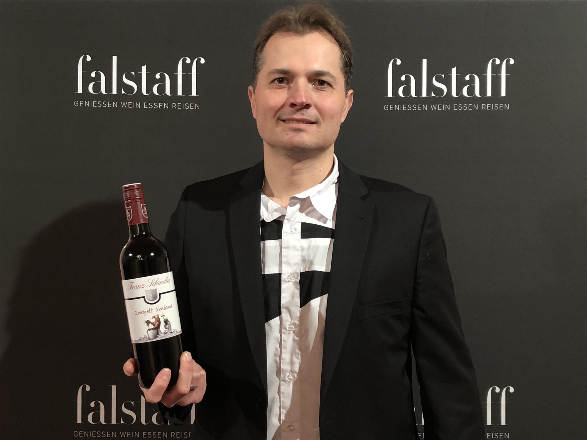 Kellermeister Ferry in elegantem Anzug hält stolz eine Flasche Wein vor einem 'falstaff' Logo.