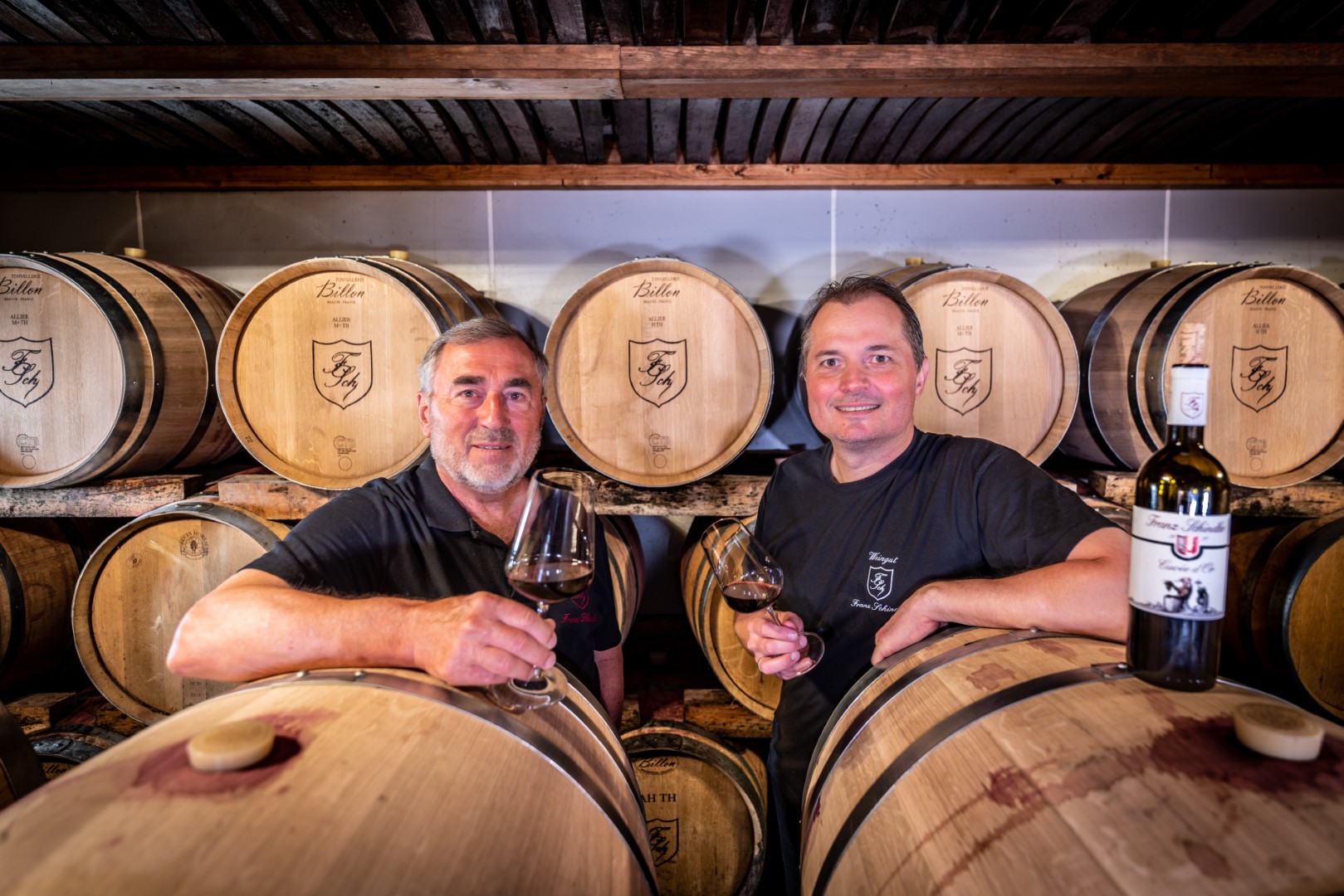 Zwei lächelnde Männer mit Weingläsern vor einem Regal voller Weinfässer im Weinkeller, Stolz auf ihre Arbeit ausstrahlend.