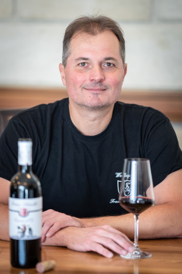 Ferry Schindler in einem schwarzen T-Shirt des Weinguts sitzt an einem Tisch, neben ihm eine Flasche Rotwein und ein gefülltes Glas.