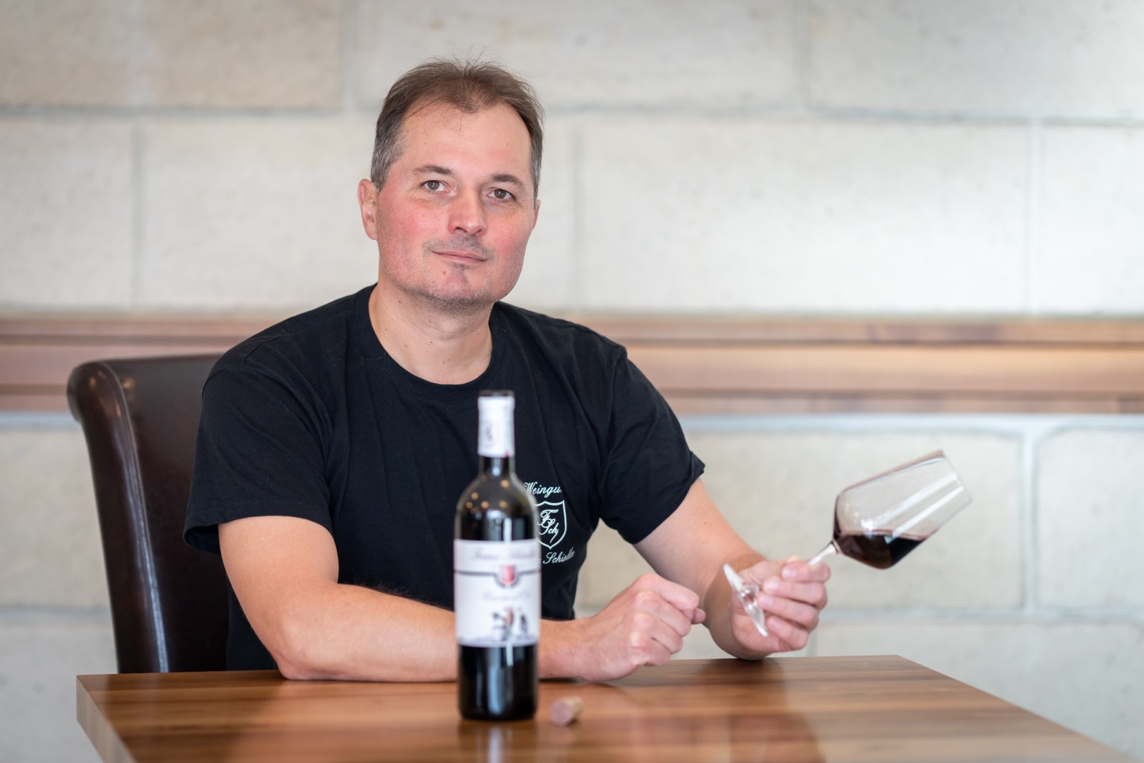 Kellermeister Ferry sitzt an einem Holztisch im Weingut, mit einem Glas Rotwein in der Hand und einer Flasche Wein vor sich.