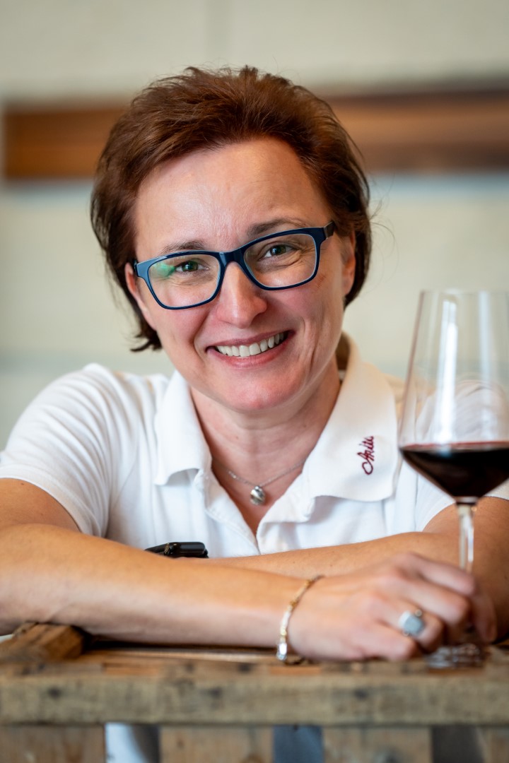 Eine fröhliche Frau im Polohemd mit Weingut-Logo lehnt an einem Holzfass, ein Glas Rotwein in der Hand.