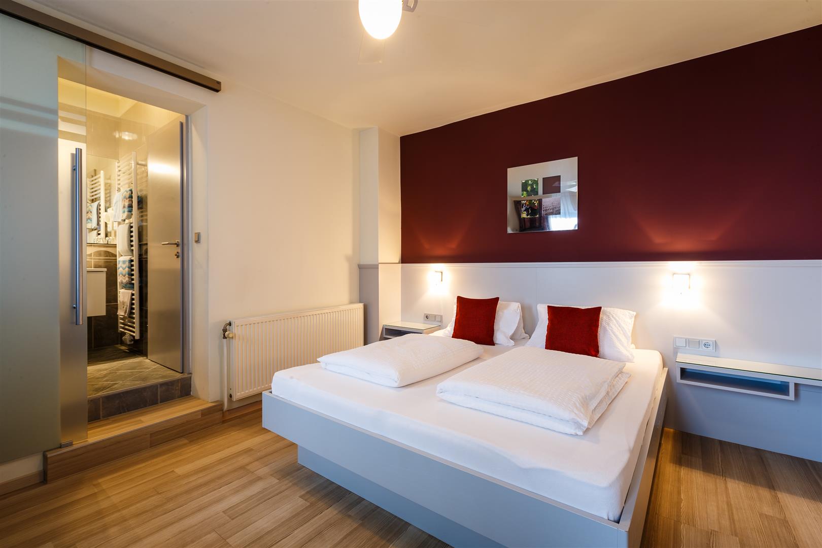 Modern eingerichtetes Gästezimmer mit weißem Bett und roten Akzentkissen, offener Badezimmertür im Hintergrund.